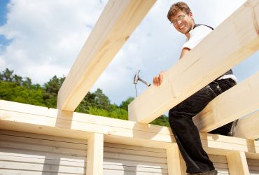 Happy carpenter sitting on the the roof beams and hammering a large nail
